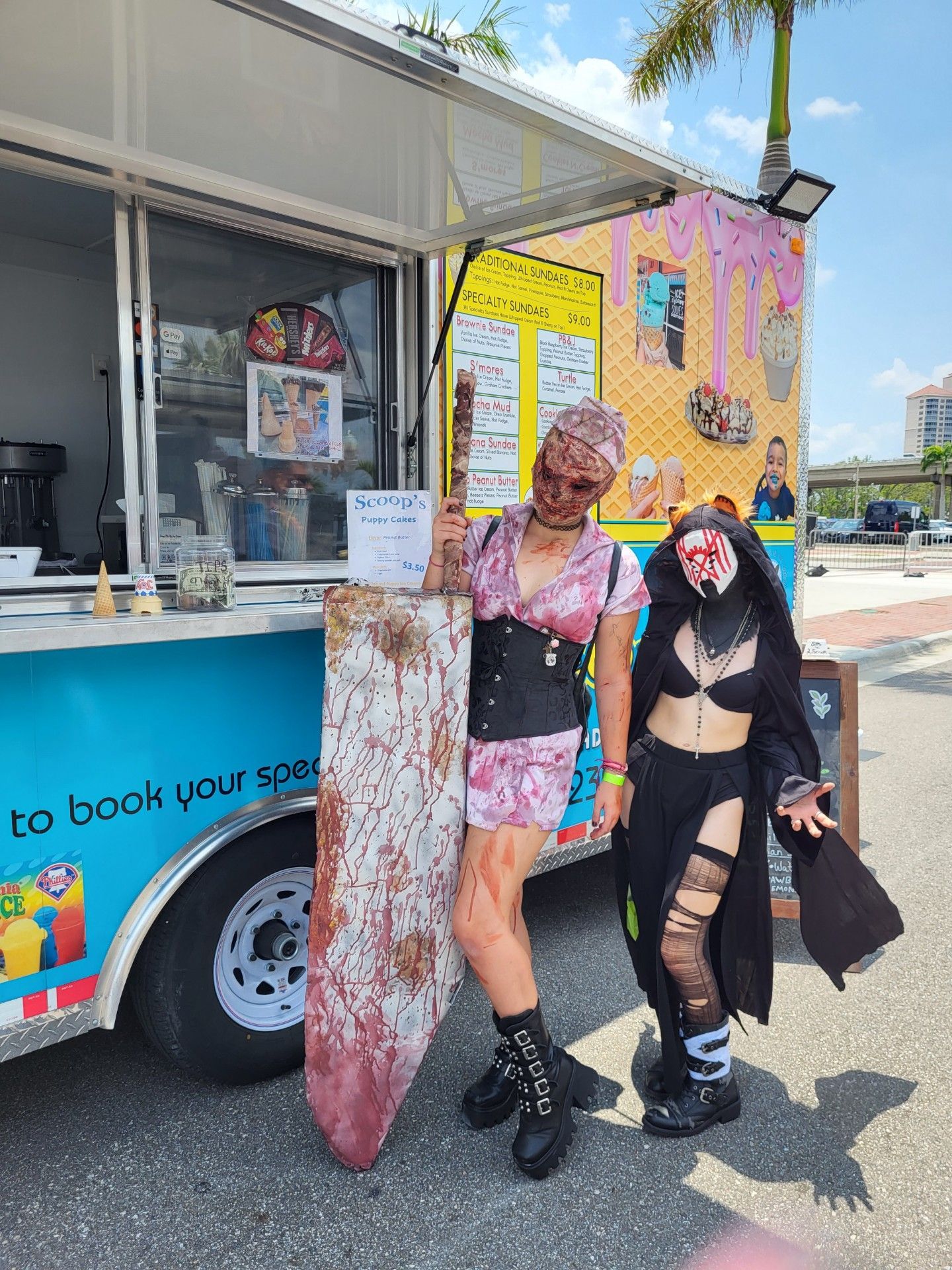 Two people in costume pose by an ice cream truck. One holds a bloody prop, the other wears a mask and dark attire.