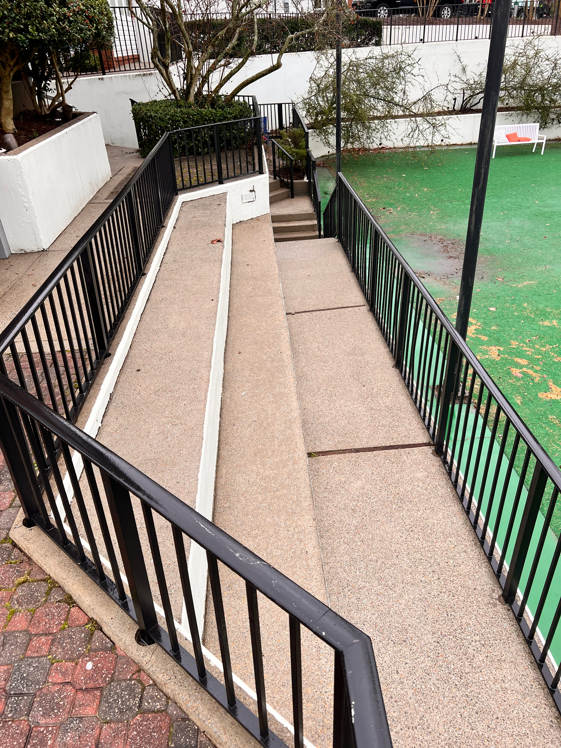 Stairway with black railings descends to green turf, flanked by white walls and a brick walkway.