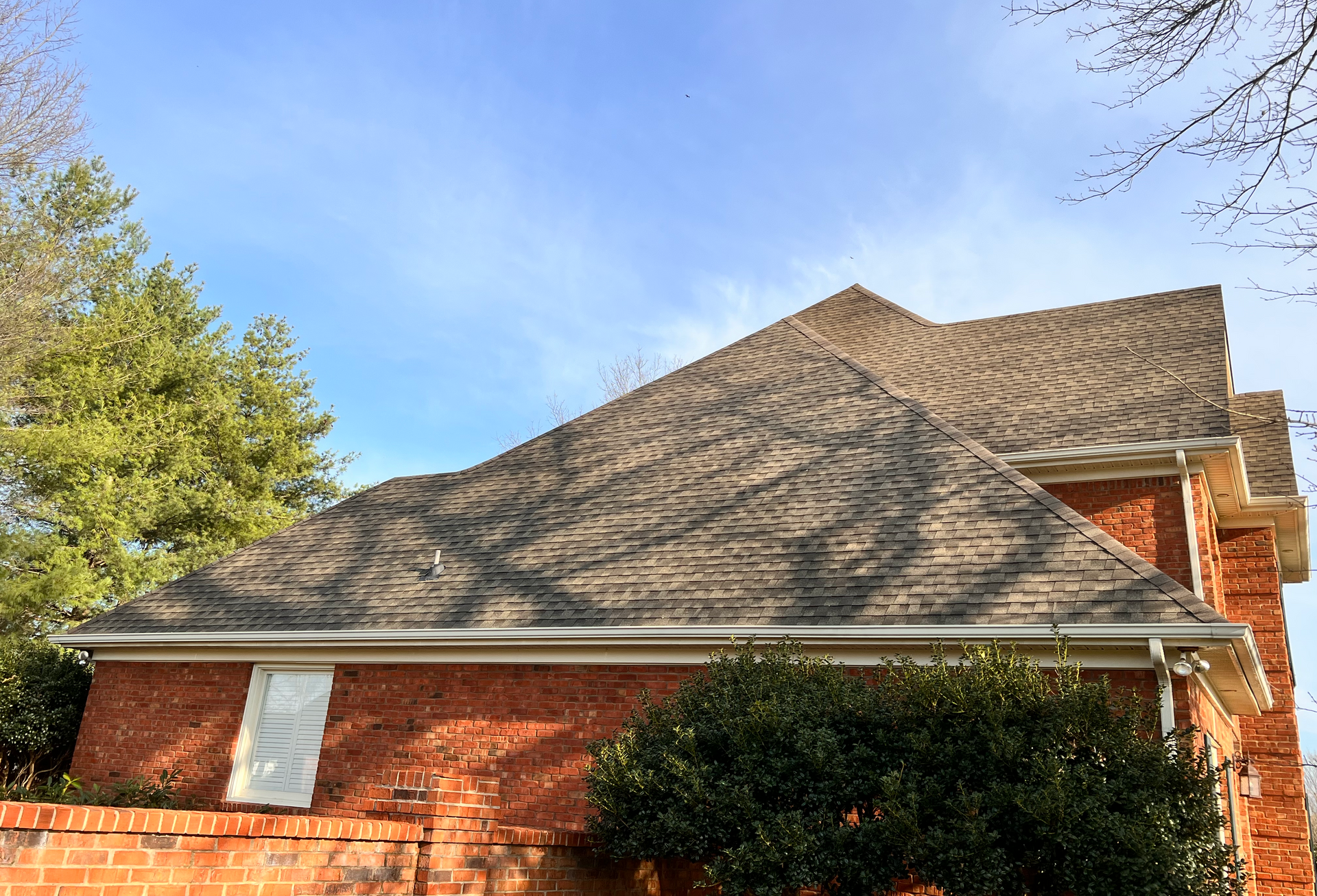 Brick house with brown shingle roof against a blue sky, trees in the background.