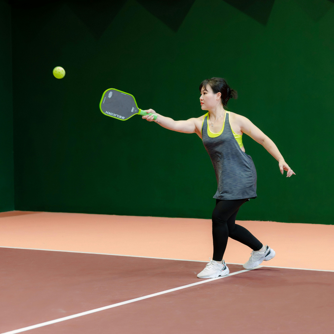 Woman playing pickleball, reaching for ball, green paddle, dark court, green wall backdrop.