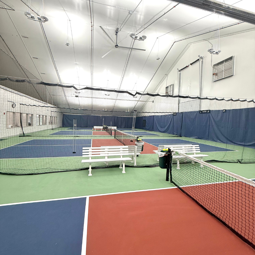 Indoor tennis court with blue and green flooring, nets, benches, and overhead lights