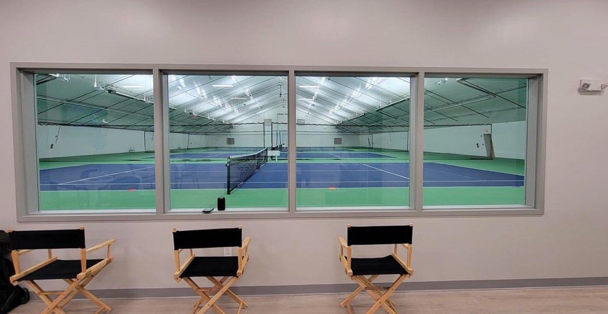 Indoor sports facility with a view of a green and blue field through a large window, chairs in the foreground
