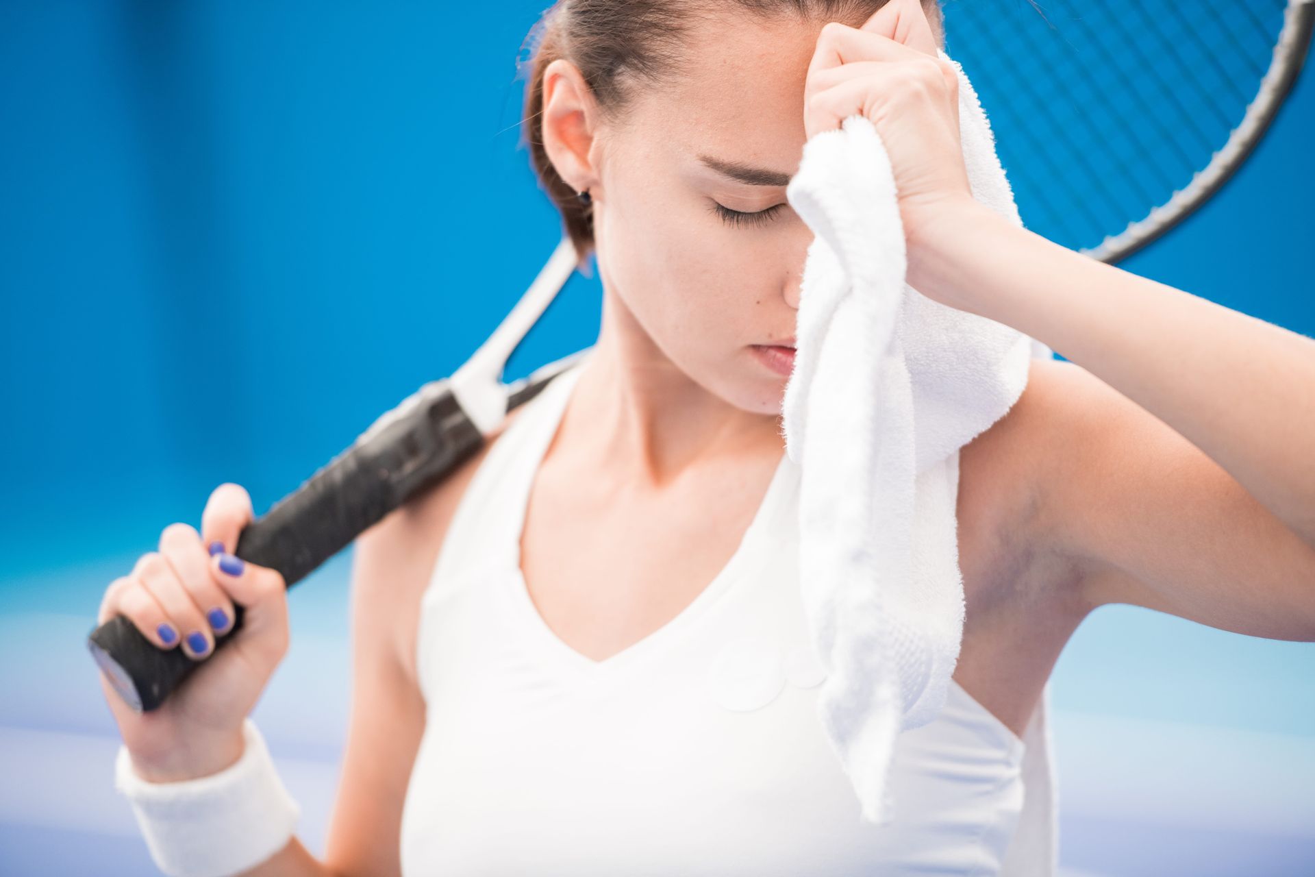 Tired tennis player wiping sweat with a towel while holding a racket on a blue court