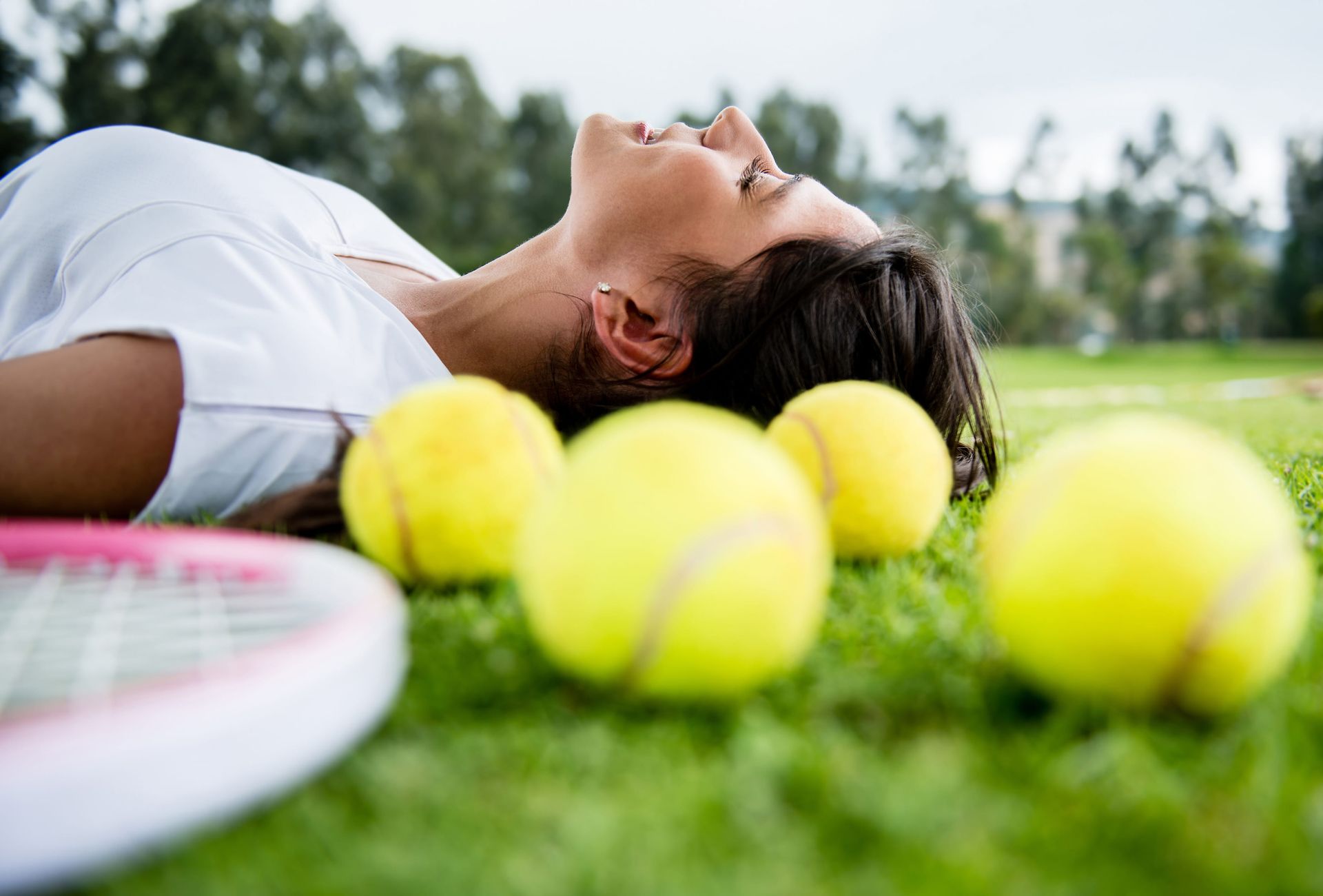 Tennis player lying on grass beside several tennis balls and a racket, resting outdoors.