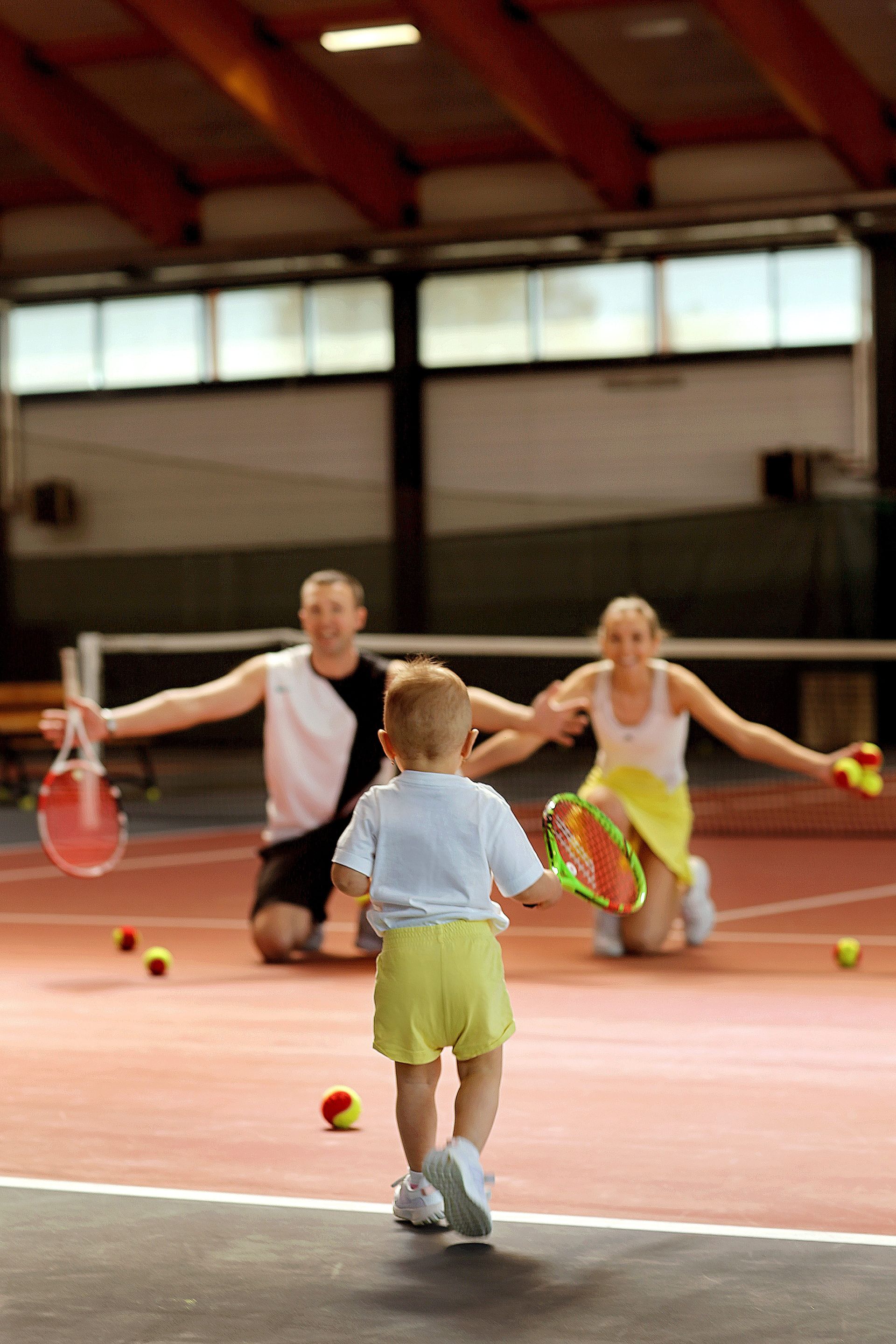 Child running toward two adults holding tennis rackets on an indoor court