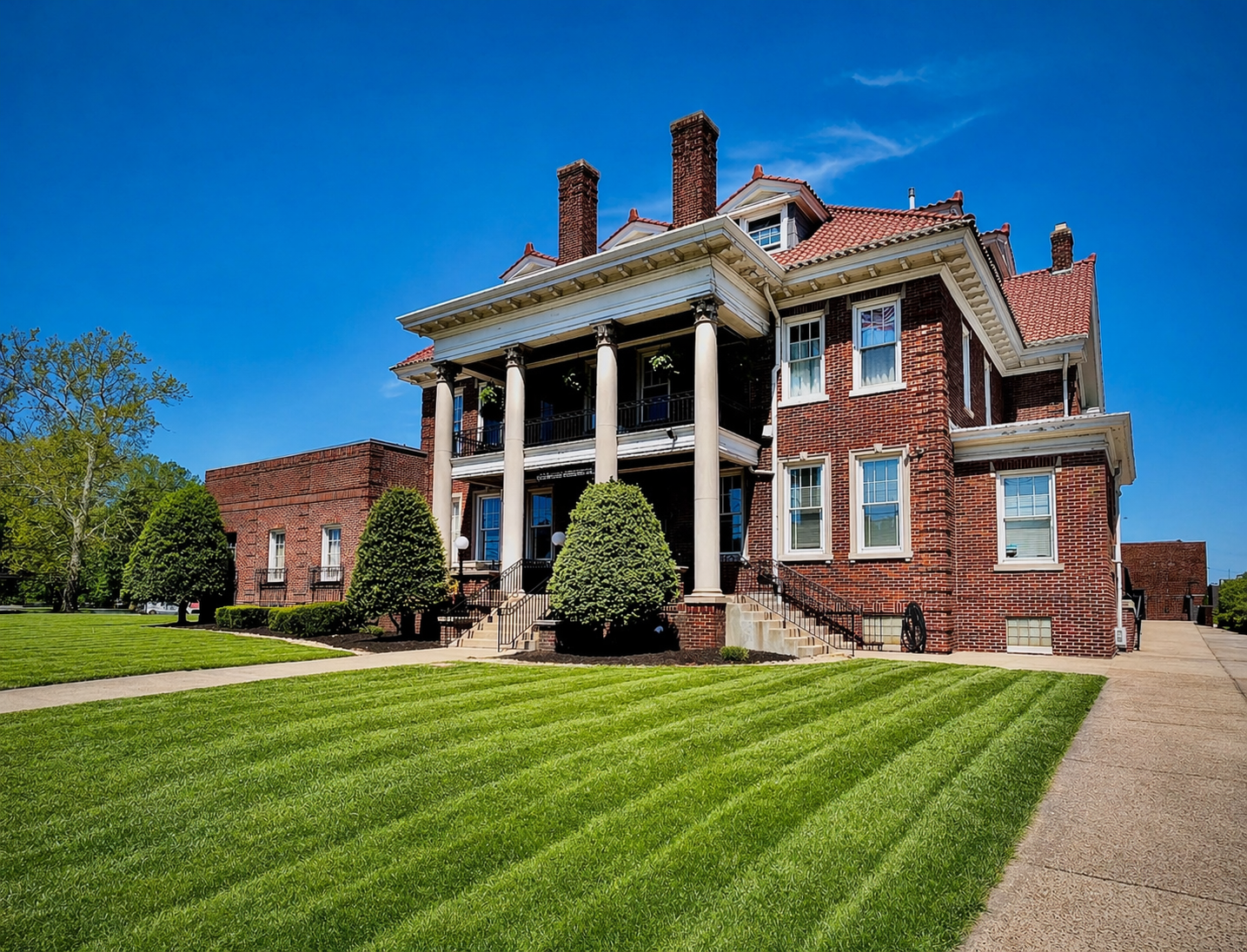 Large brick mansion with white columns, red roof, and manicured lawn on a sunny day