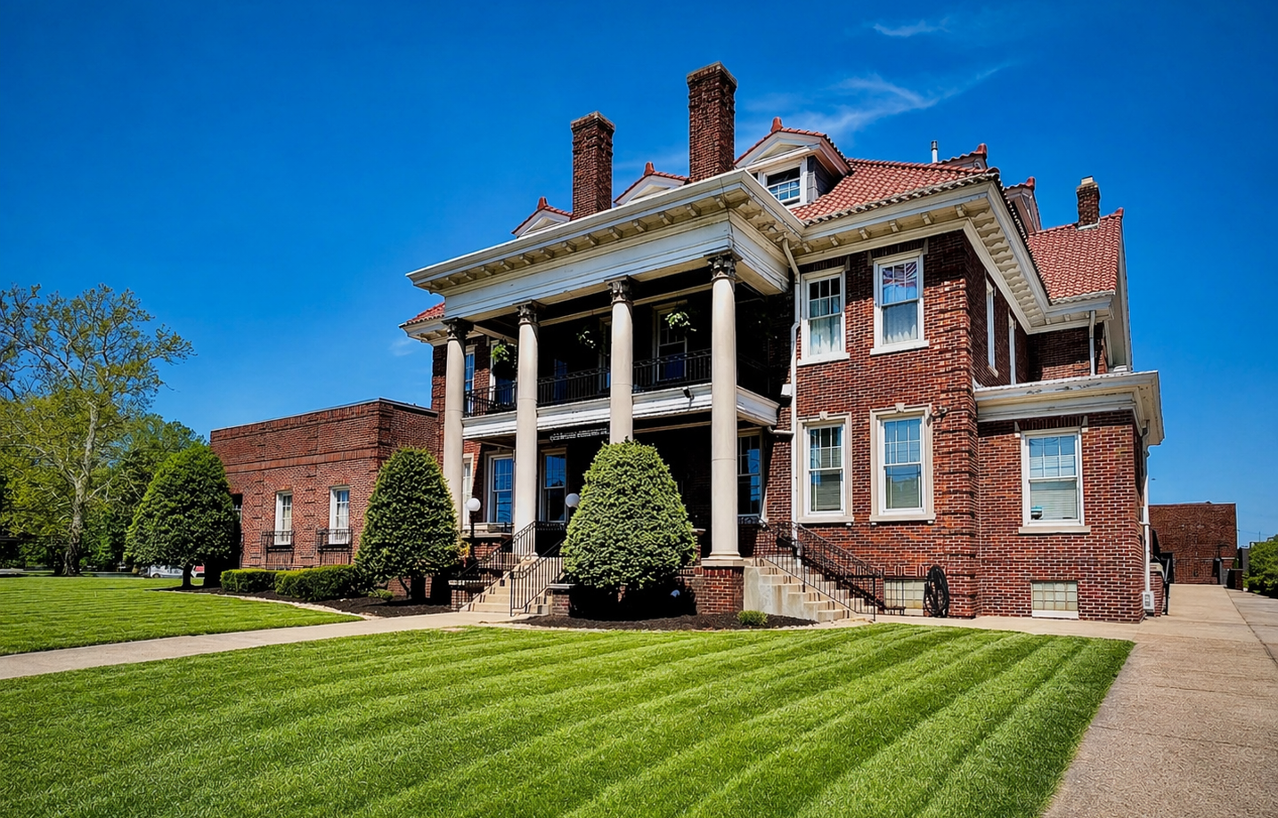Large red-brick mansion with white columns and manicured lawn under a bright blue sky