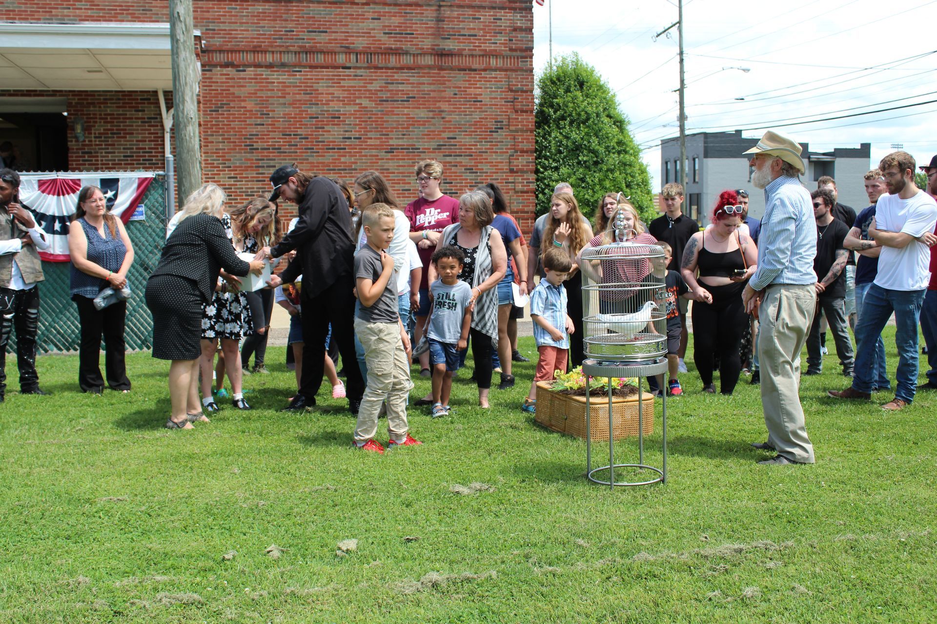 Group of people gathered on grass outside a brick building, watching a small event near a flower display.