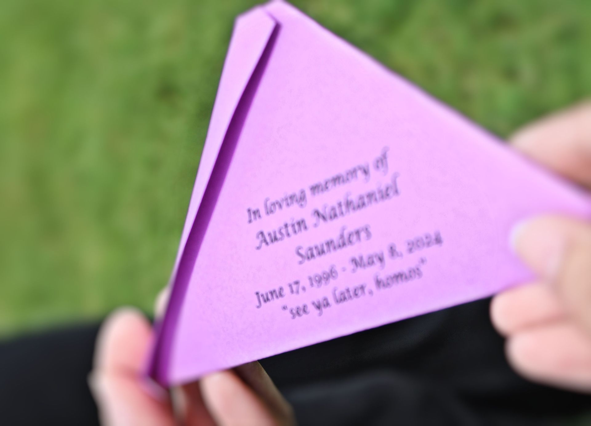 Purple folded memorial card reading “In loving memory of Austin Nathanael Saunders” held outdoors on grass.