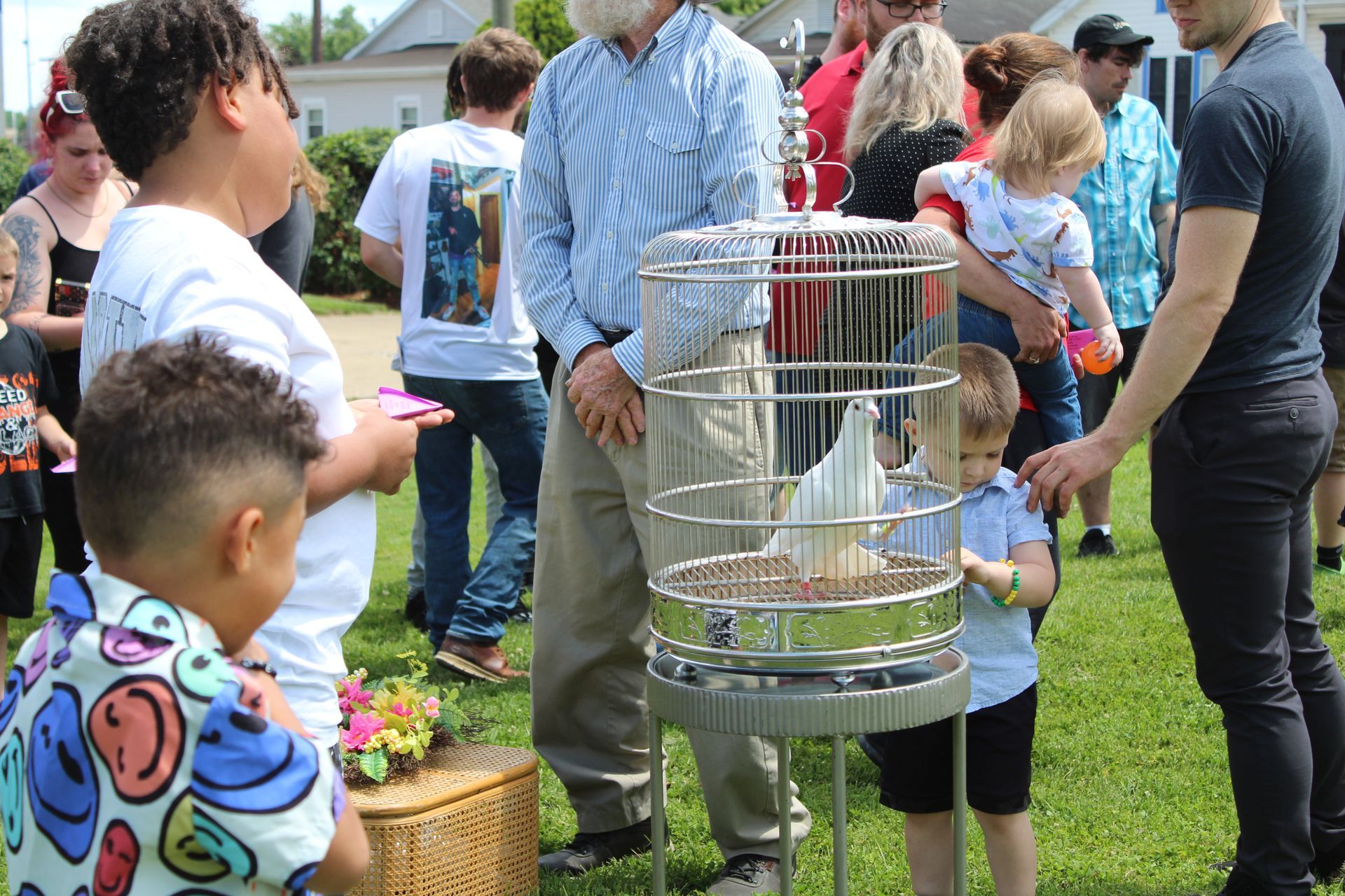 People gathered around a small birdcage outdoors during a community event, with children nearby.