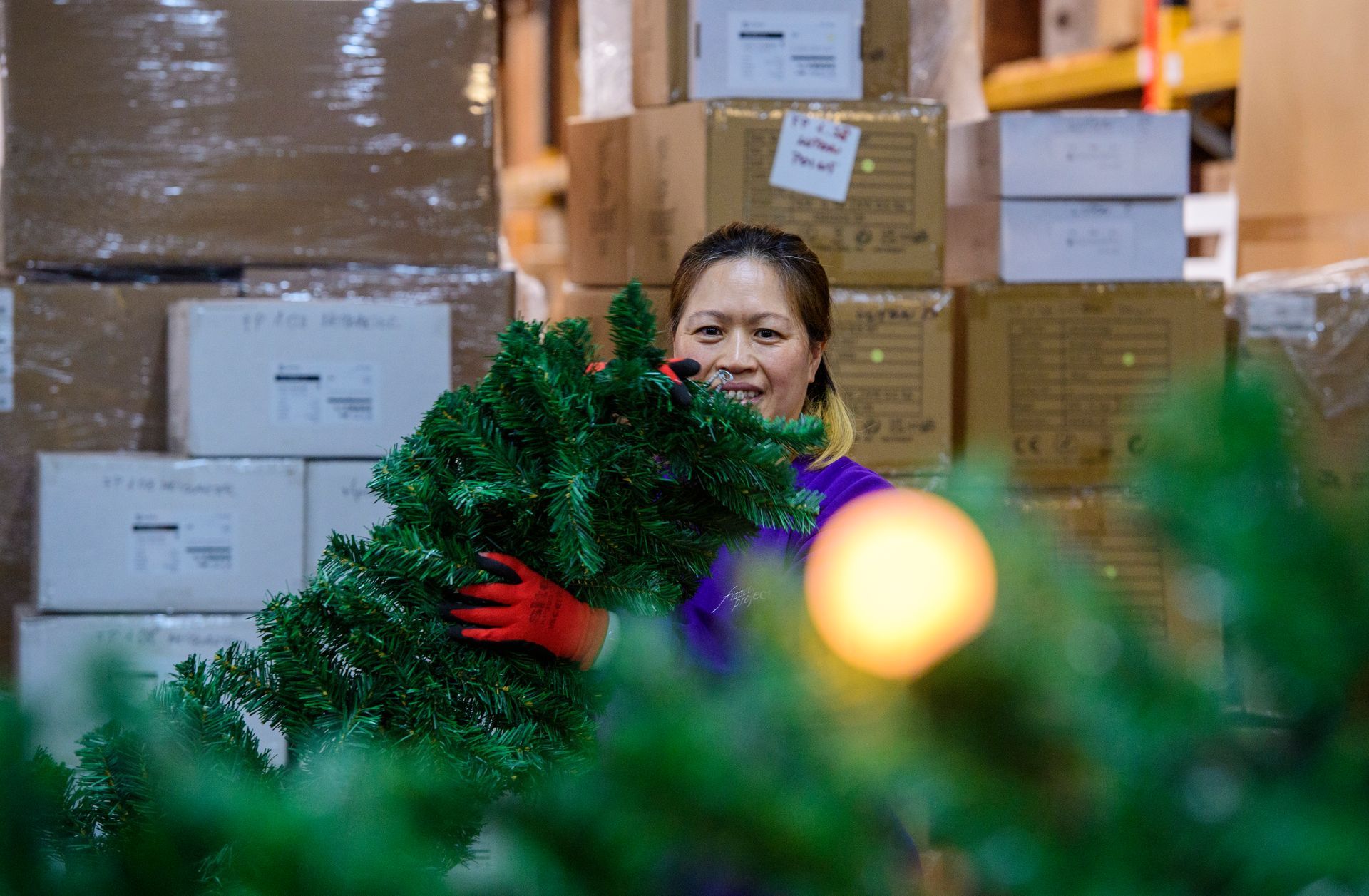 Person in red gloves arranging green wreaths among stacked boxes in a storage room