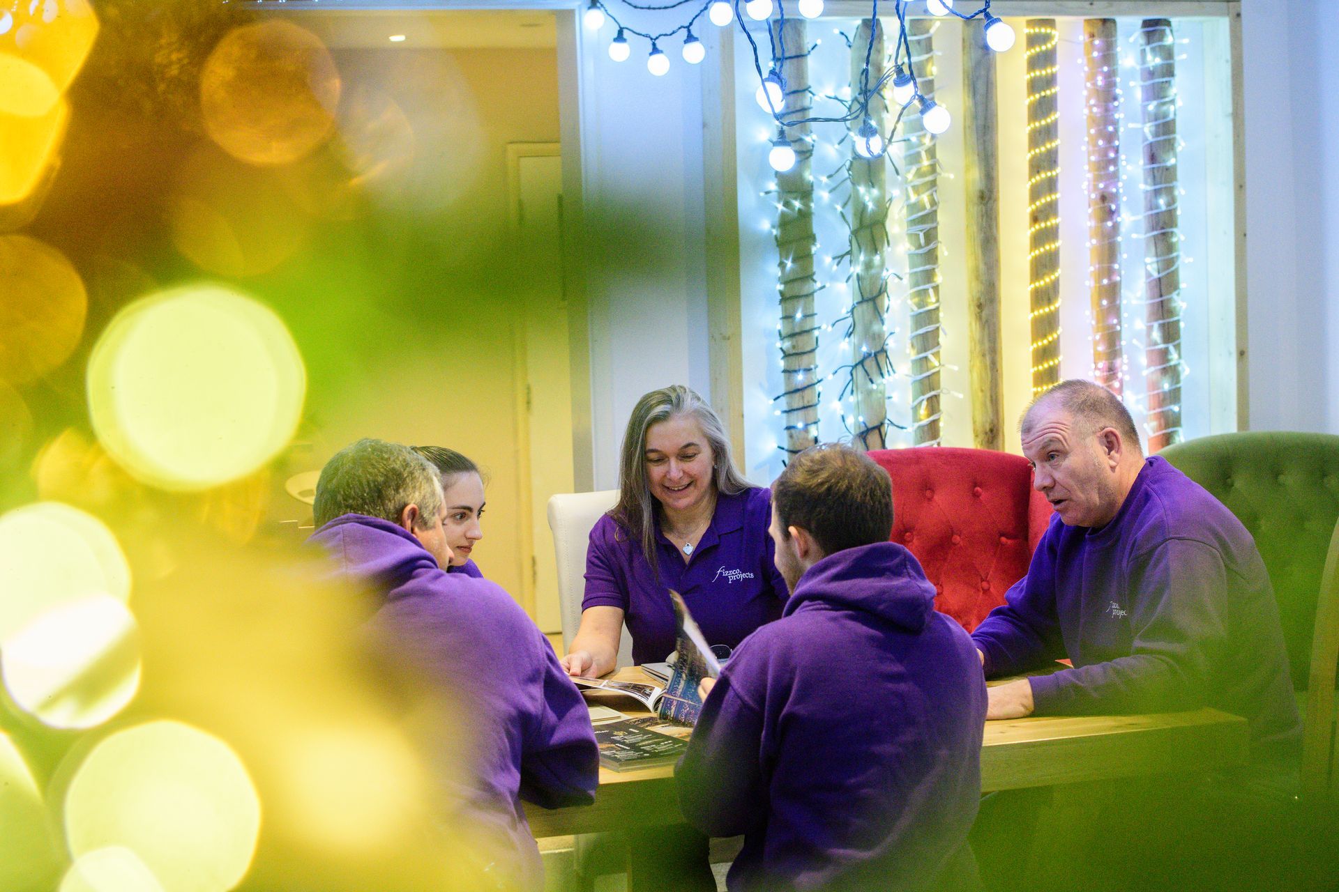 Four people in purple shirts chat at a table in a softly lit room with festive decorations.
