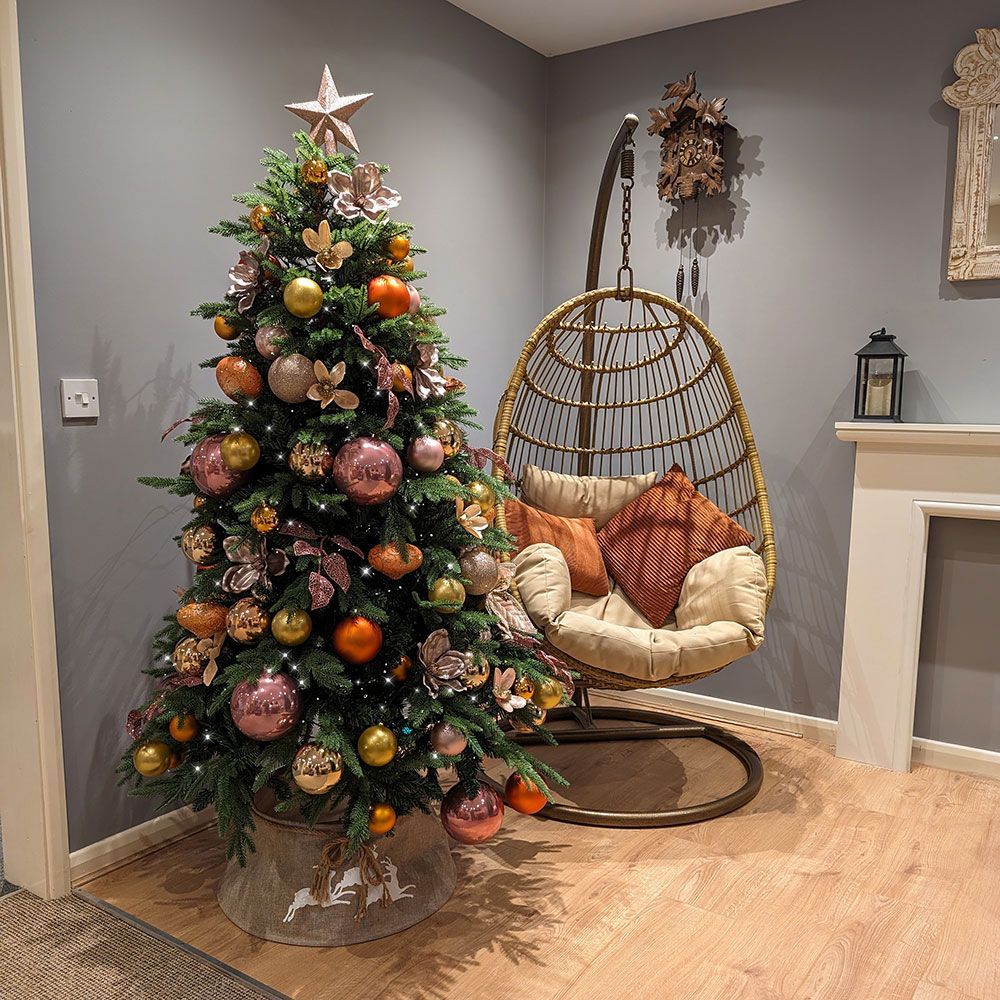 Decorated Christmas tree beside a wicker hanging chair in a cozy corner with a fireplace mantel.