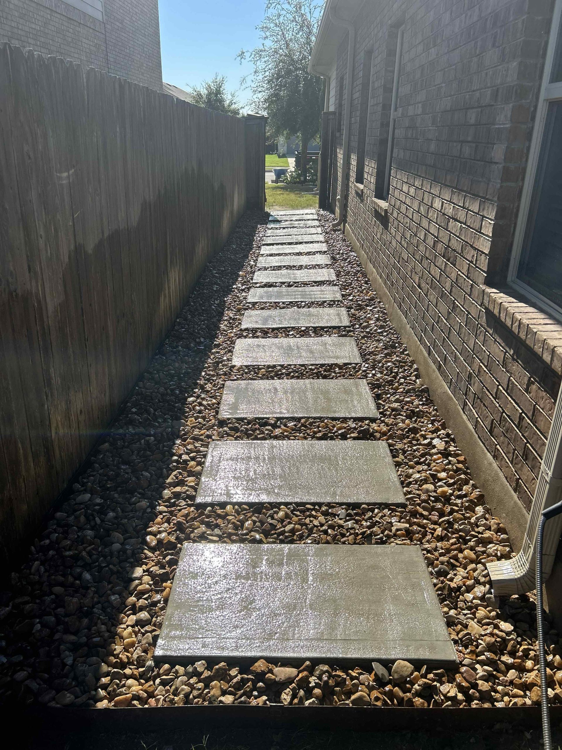 A narrow outdoor path made of large, flat grey stone pavers set in a bed of brown gravel between a brick wall and a fence.
