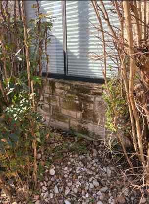 A stone foundation wall beneath a window, surrounded by bare trees and gravel mulch in a garden setting.