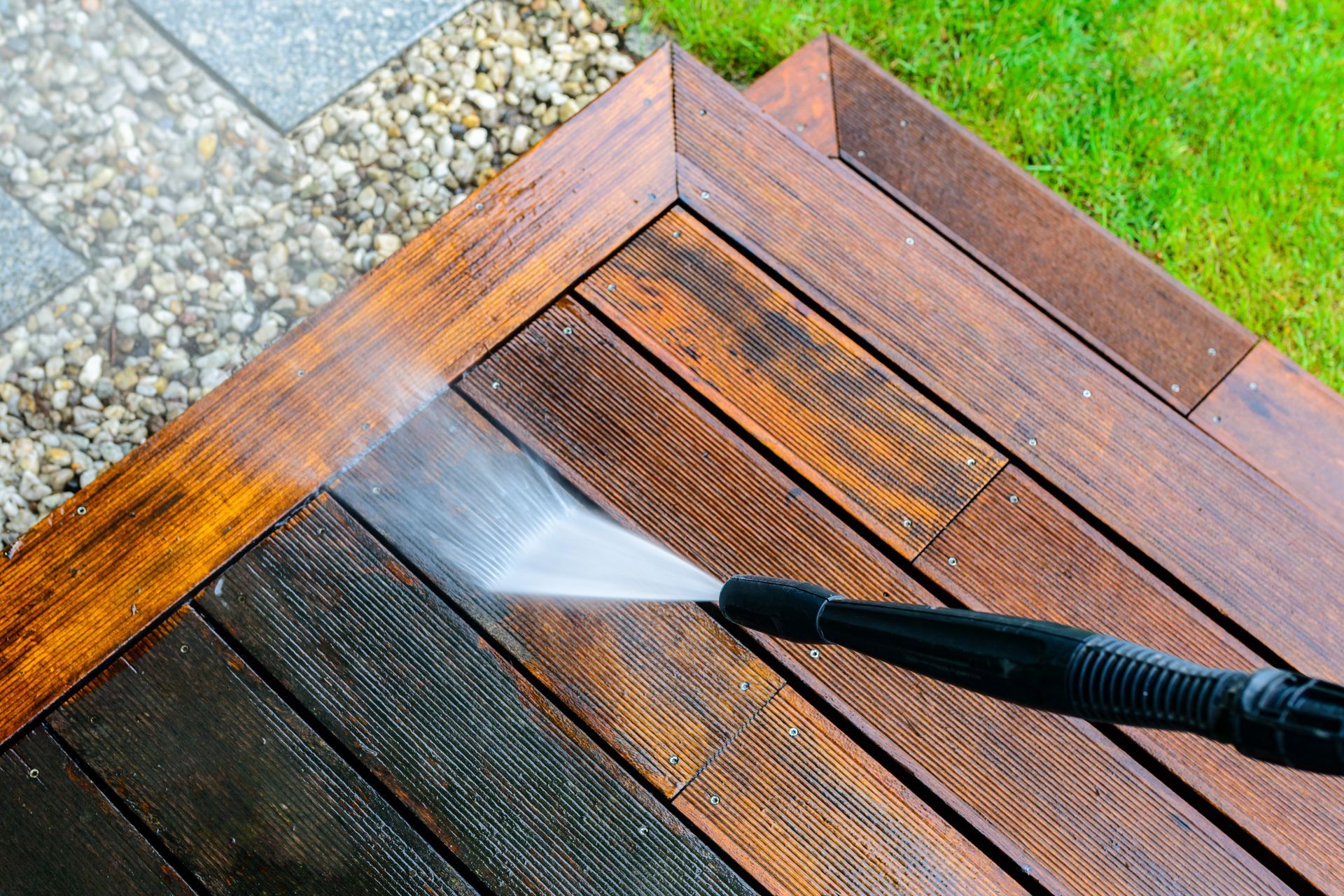 Person power washing a wooden deck, with contrast between cleaned and uncleaned sections; green lawn and shrubs visible.