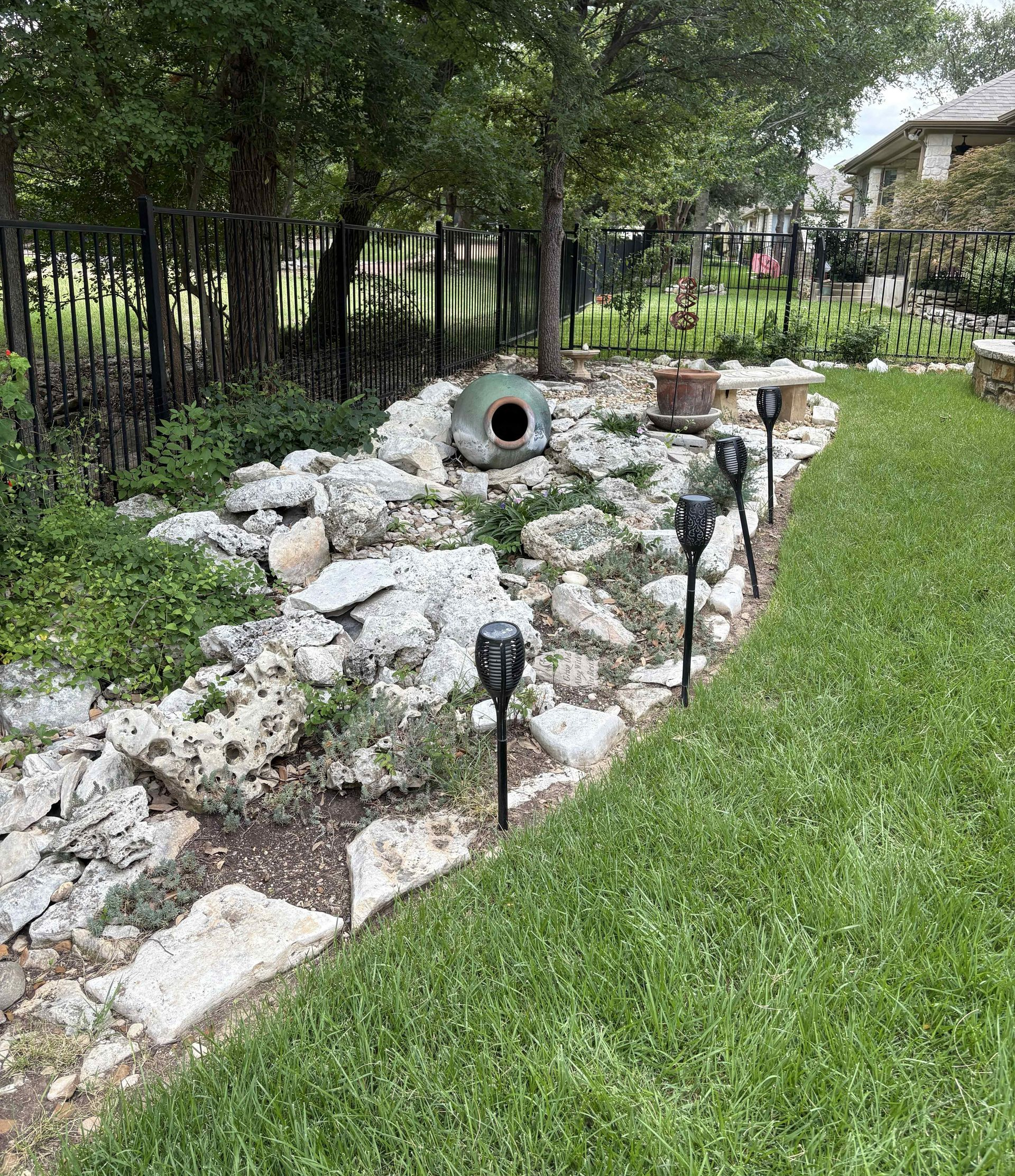 A rocky landscaping border lines a grassy lawn, featuring a ceramic jar and several black solar lights along the edge.