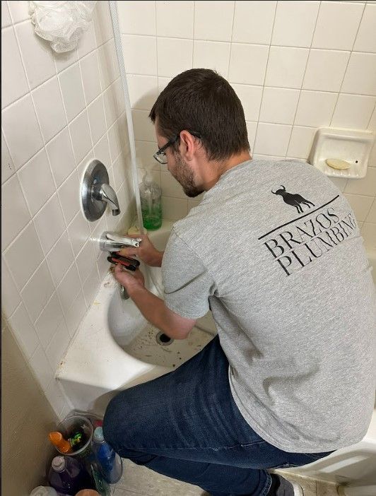 A man in a gray shirt is fixing a sink in a bathroom.