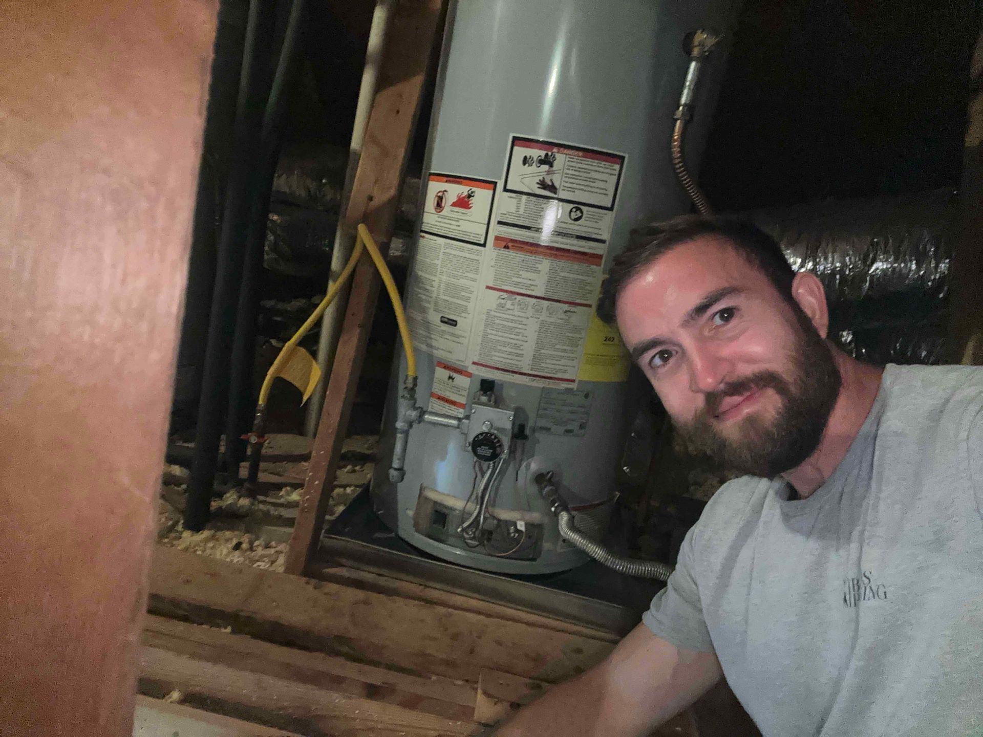 A man with a beard is standing next to a water heater in an attic.