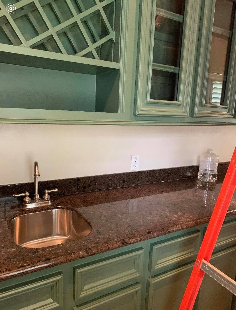 A kitchen wet bar with light green cabinets, dark granite countertops, a small stainless sink, and a red ladder.