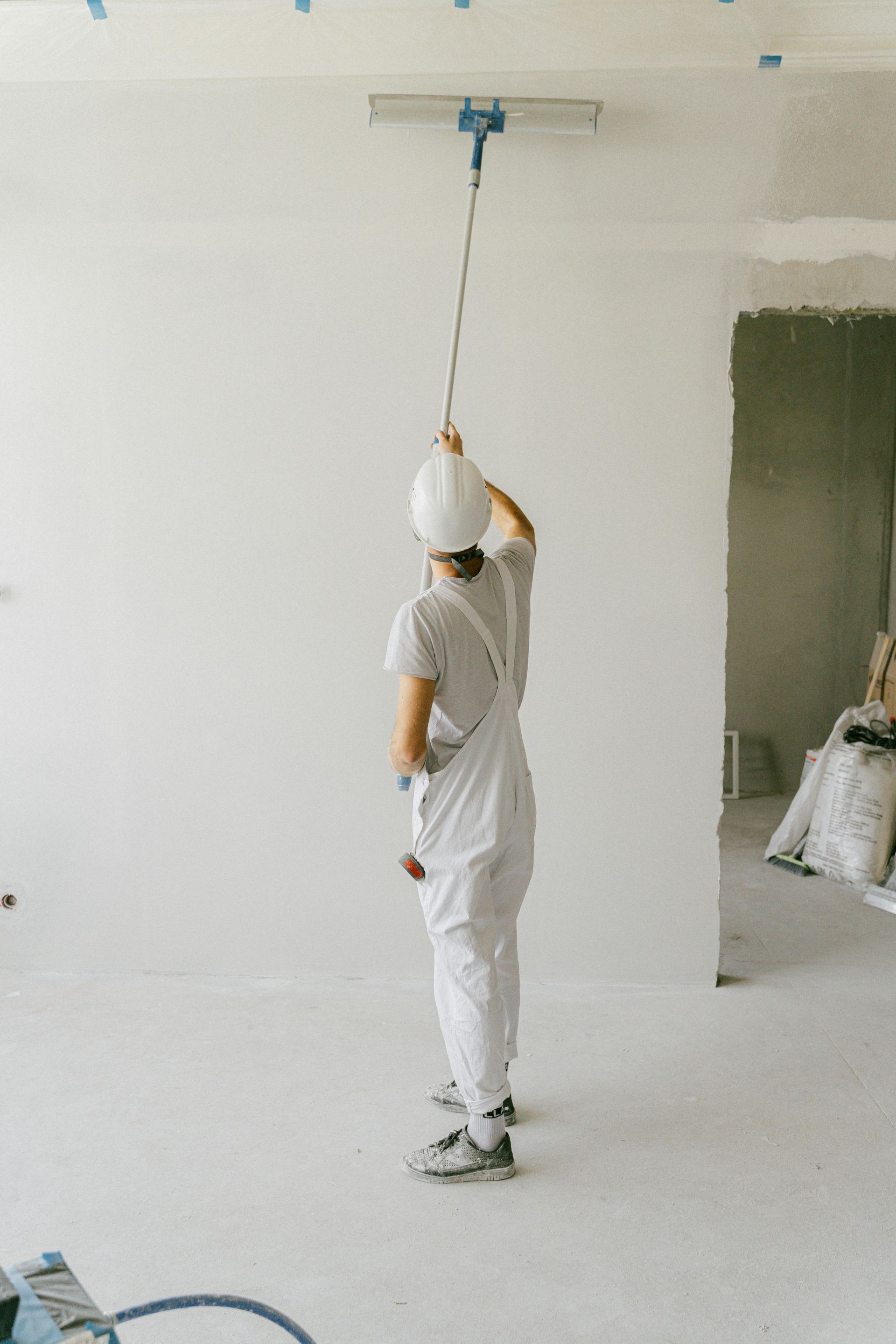 A person in white work clothes and a hard hat uses a long-handled tool to sand or smooth a white interior wall.