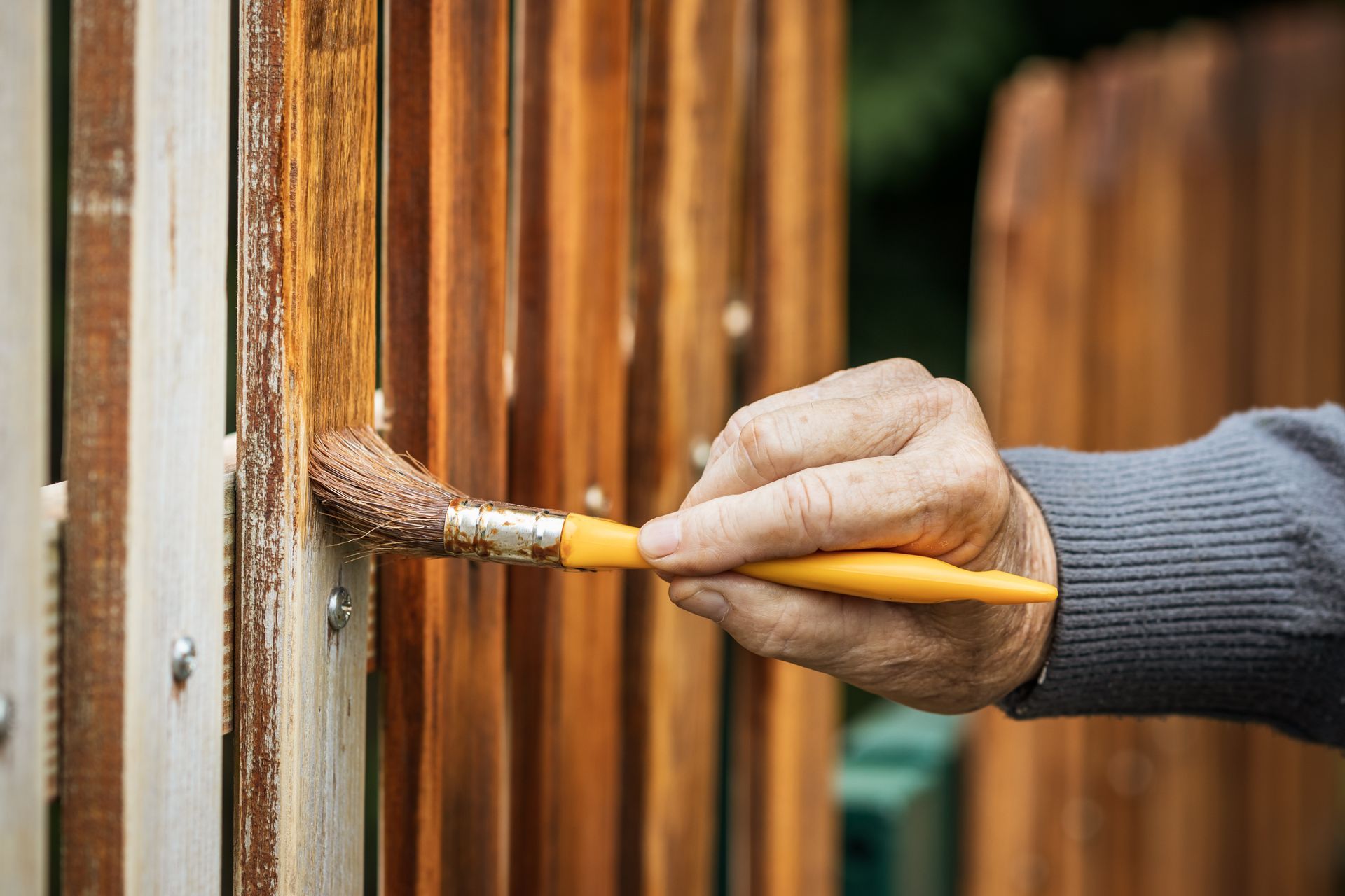 A close-up of a hand using a paintbrush to apply brown wood stain to a wooden fence.