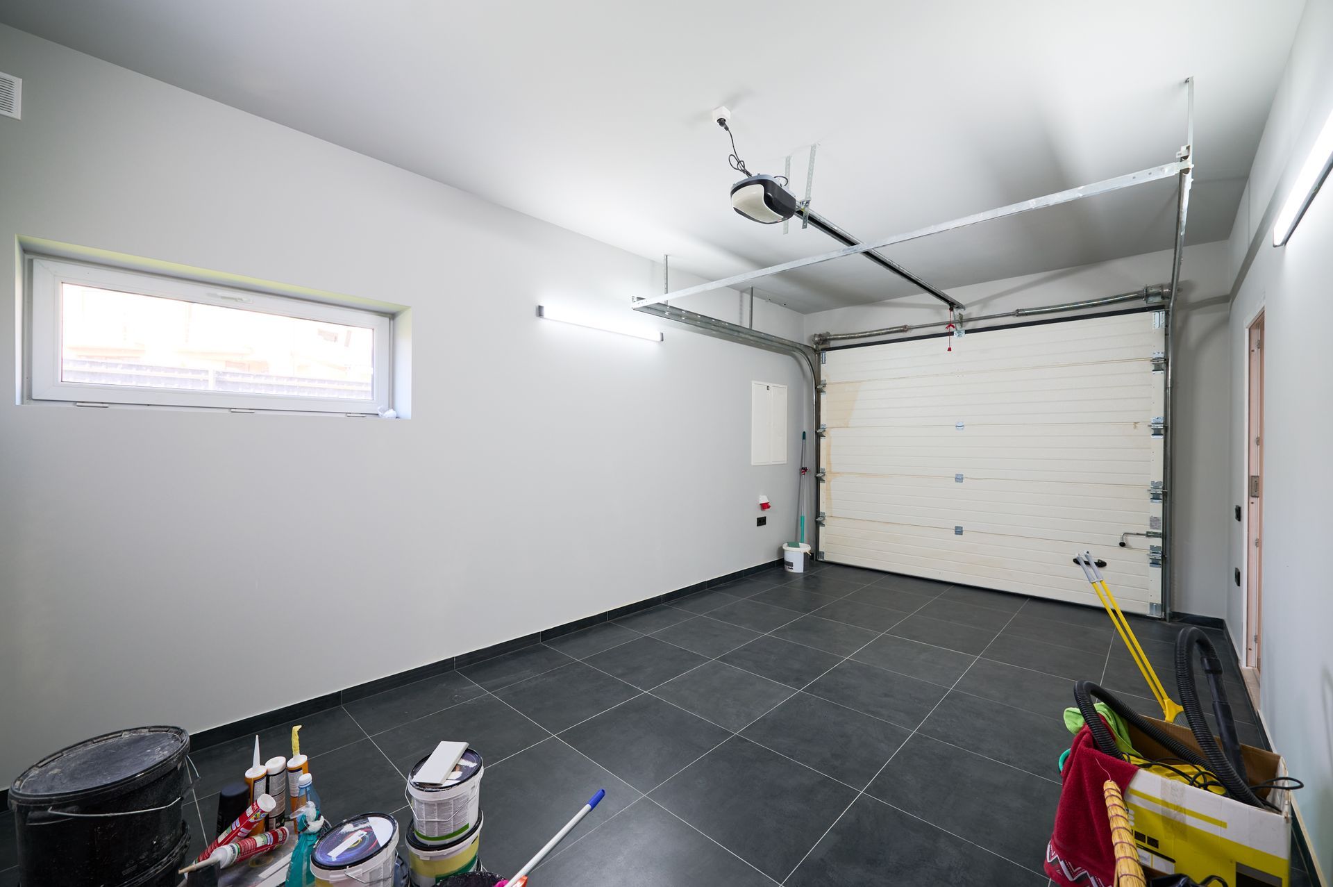 Empty white garage with black floor tiles, closed overhead door, and a few cleaning supplies in the corner