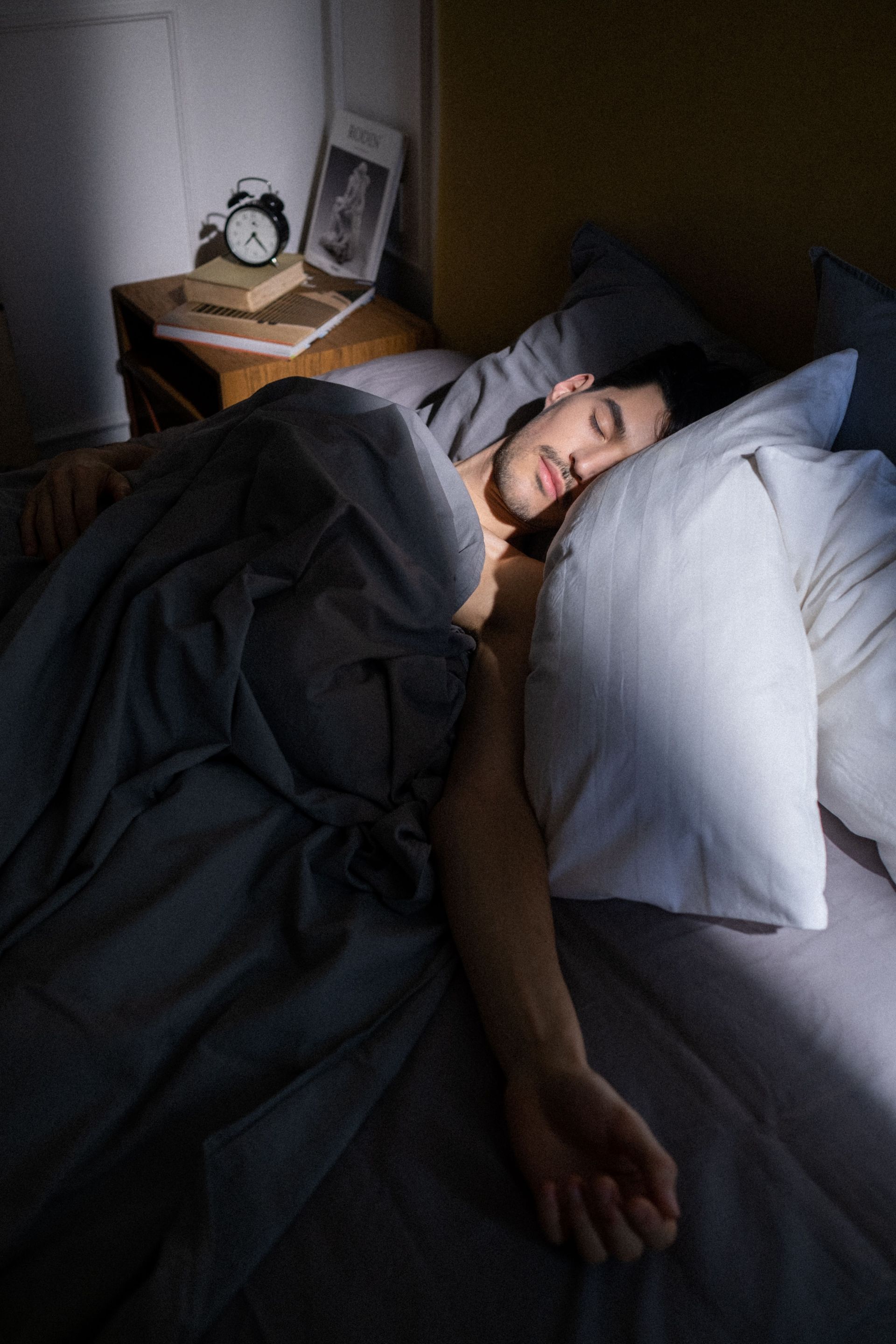 Person sleeping in bed under dark blankets, with a bedside clock and framed photo nearby.