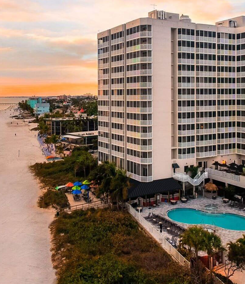 Beachfront high-rise hotel, white facade, next to beach with pool and sunset sky.