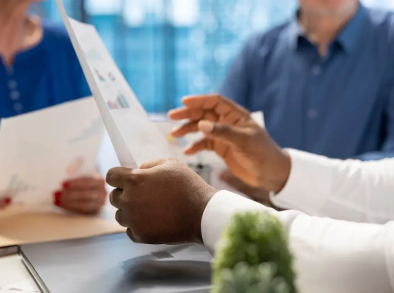 Close-up of three people at a table reviewing documents. Black hand holds paper; others' hands are visible.