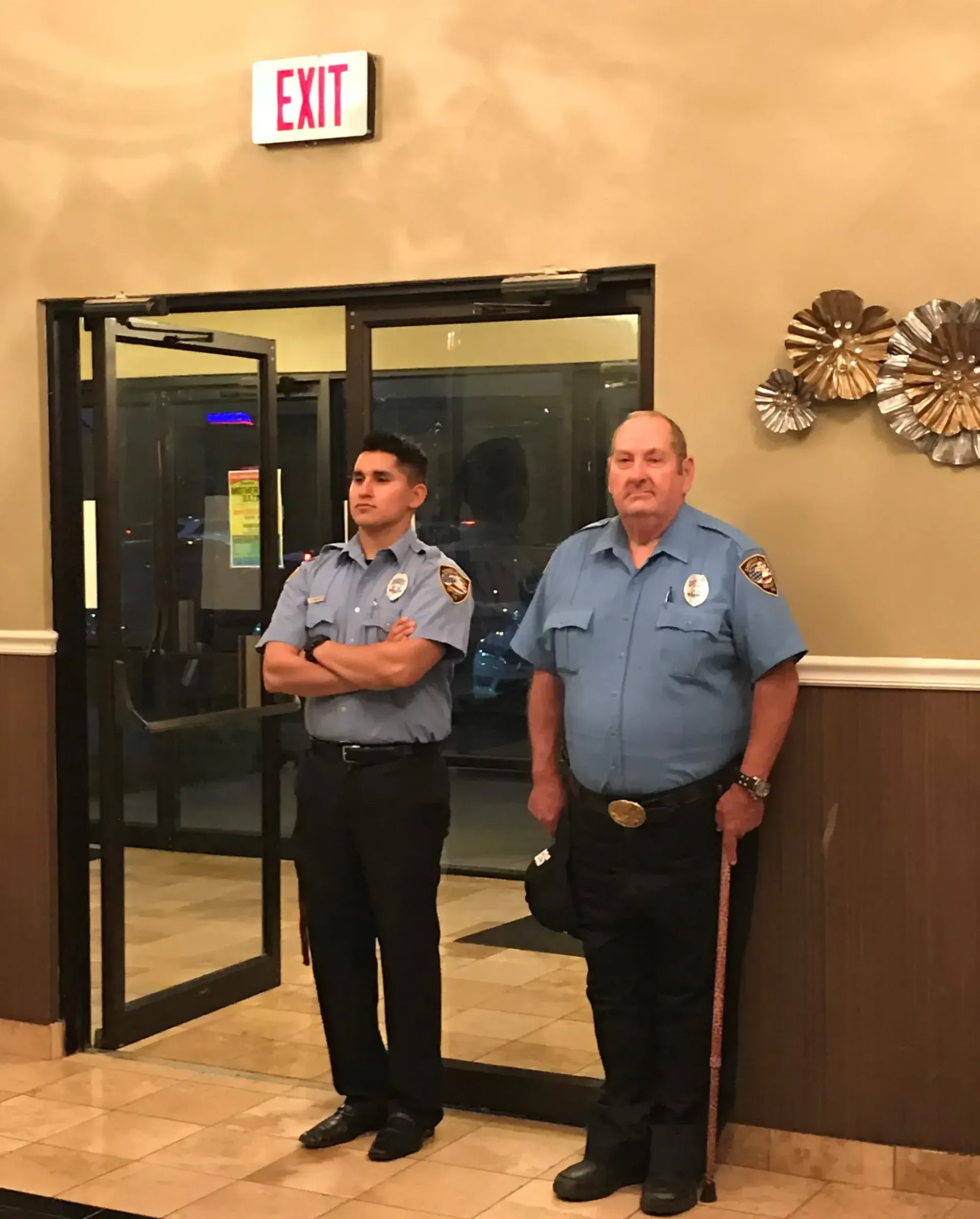 Two security guards stand by an open door under an EXIT sign in a lobby.