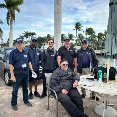 Six men posing outdoors by a palm tree and table on a breezy patio with parked cars nearby