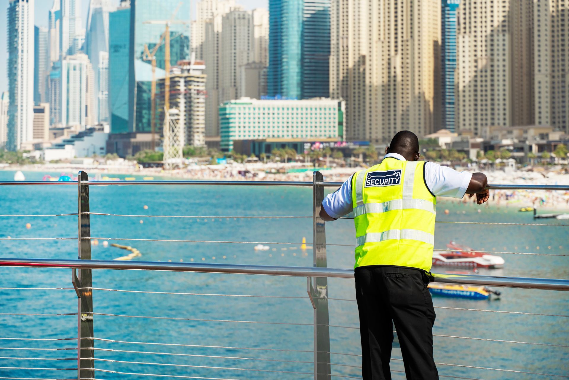 Person in a neon safety vest stands by a waterfront railing with a city skyline in the background.