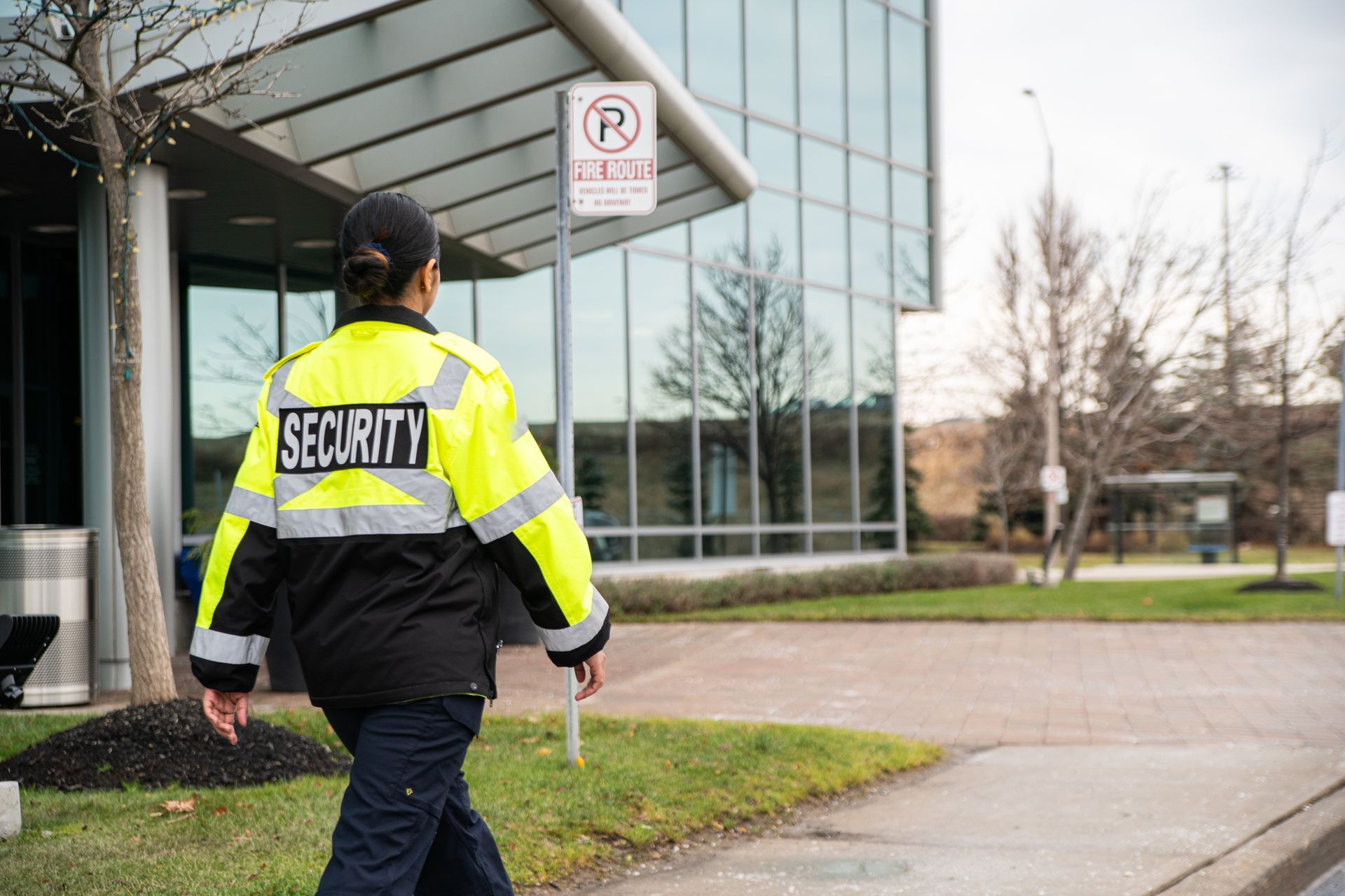 Security guard in a yellow jacket walking outside a glass office building near a no-parking sign