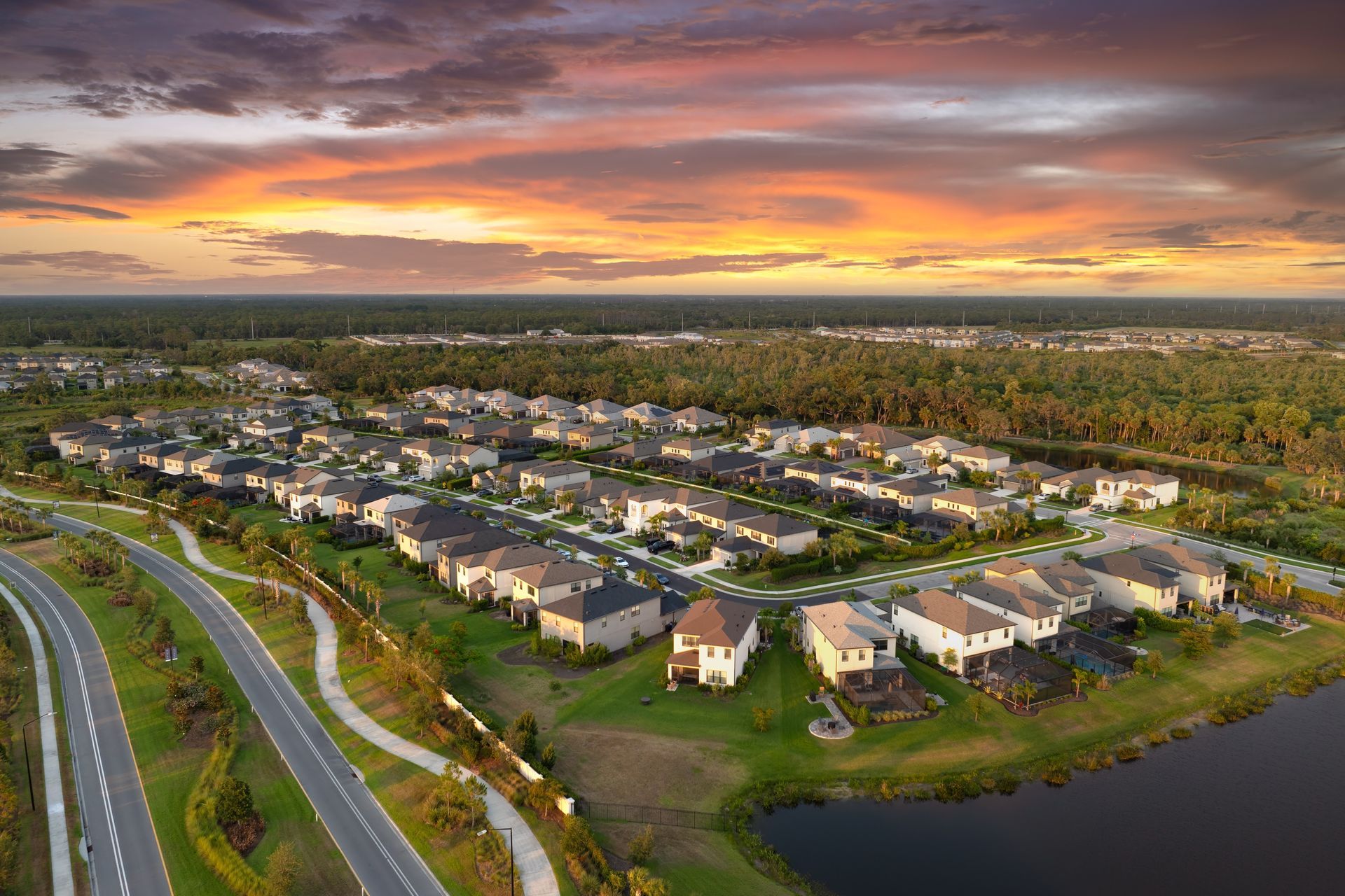 Aerial view of a suburban neighborhood beside a lake at sunset, with orange-pink skies and winding roads