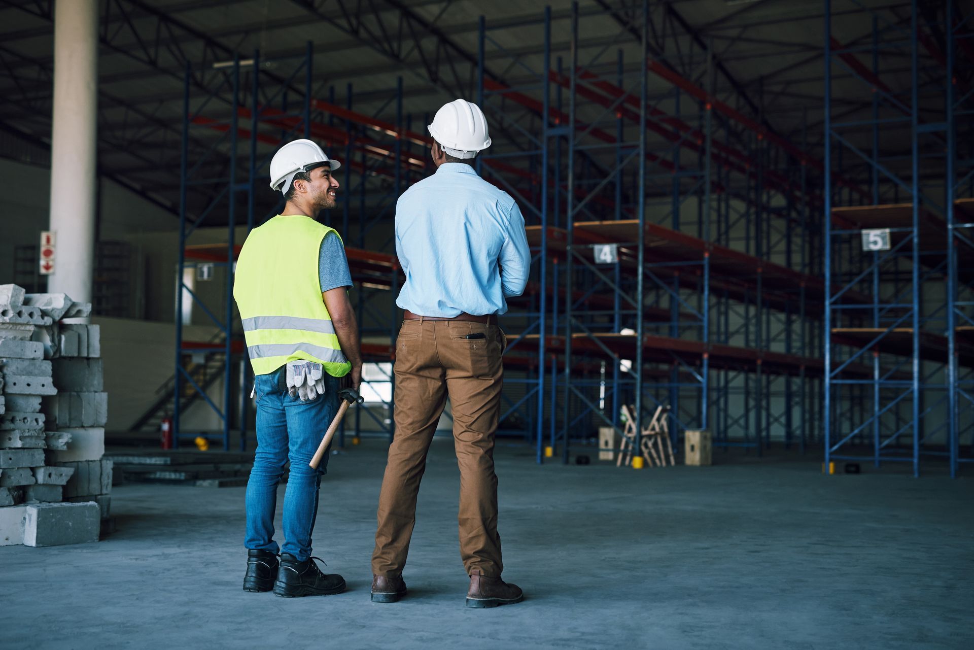 Two workers in hard hats inspect a warehouse interior with empty shelving racks.