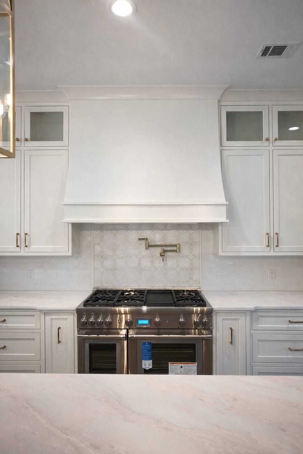 Bright white kitchen with stainless stove, marble island, and tall cabinets under a windowed hood.