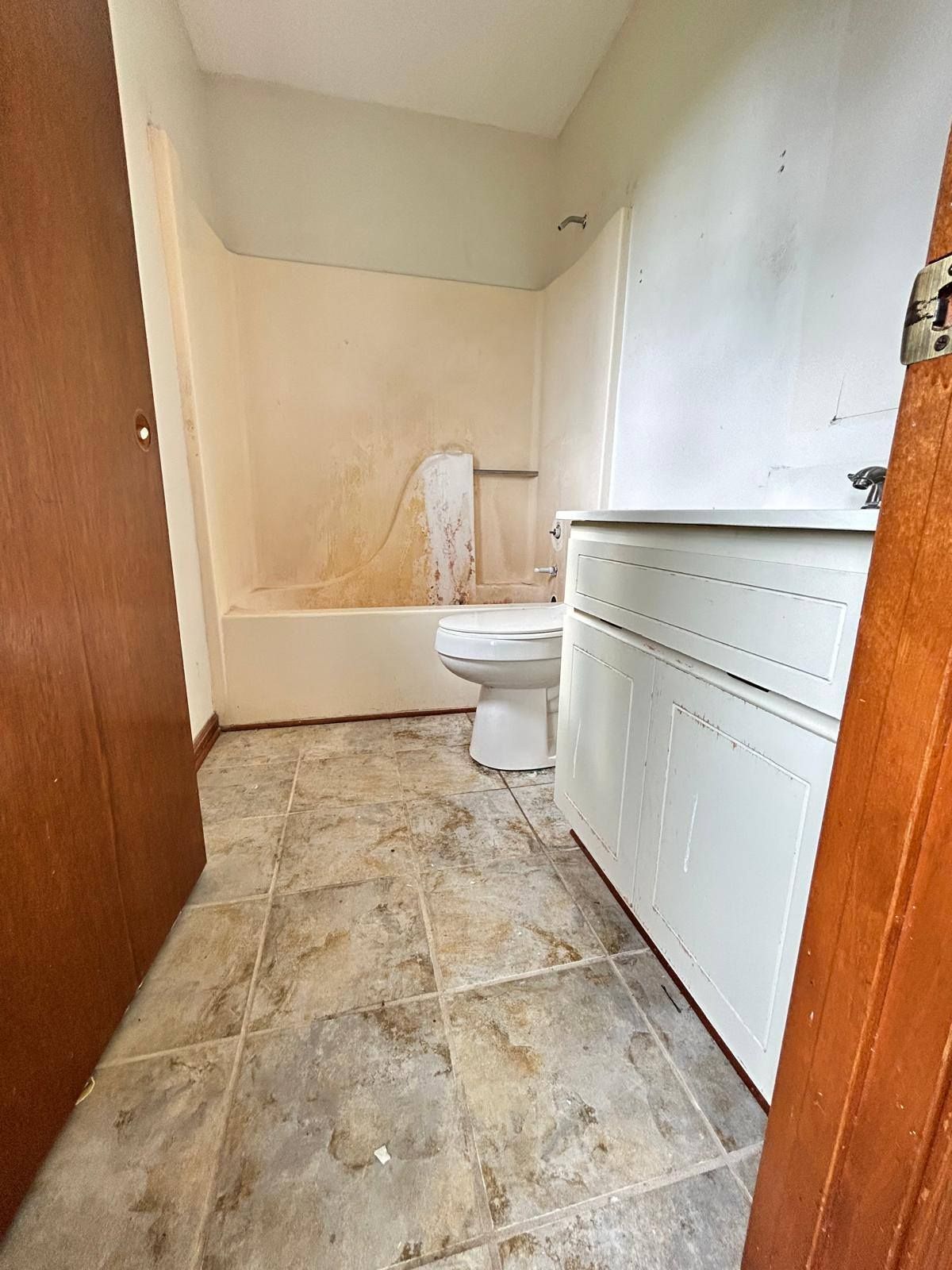 Small bathroom with a bathtub, toilet, and white vanity, showing worn beige walls and tiled floor.