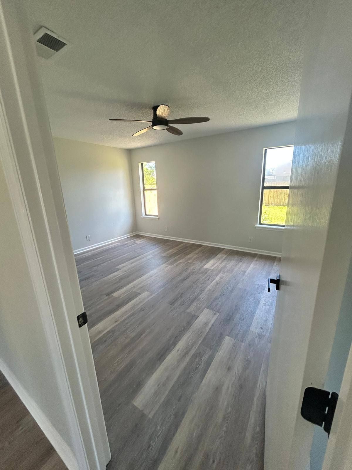 Empty bedroom with gray wood floors, white walls, ceiling fan, and two narrow windows.