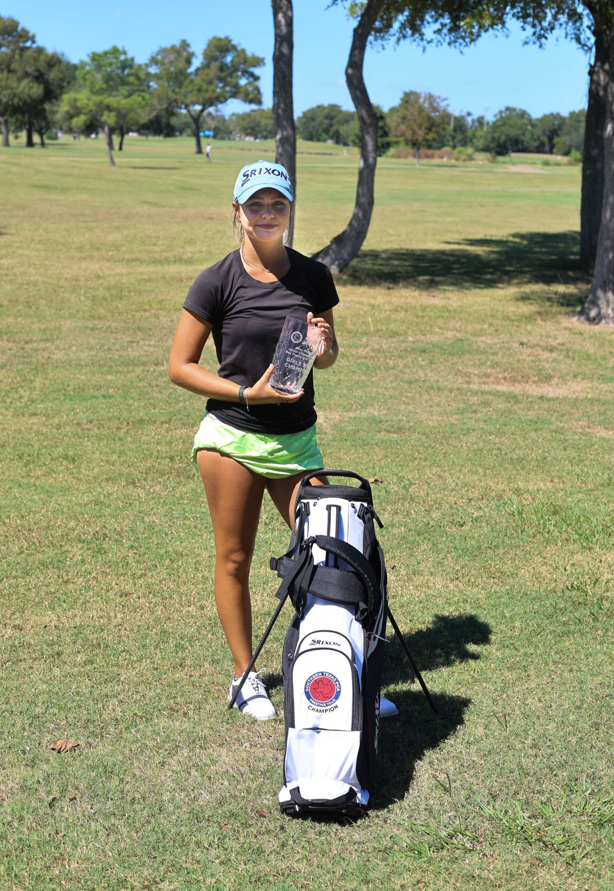 Young woman golfer in pink shirt and black shorts stands next to golf bag on a green course.