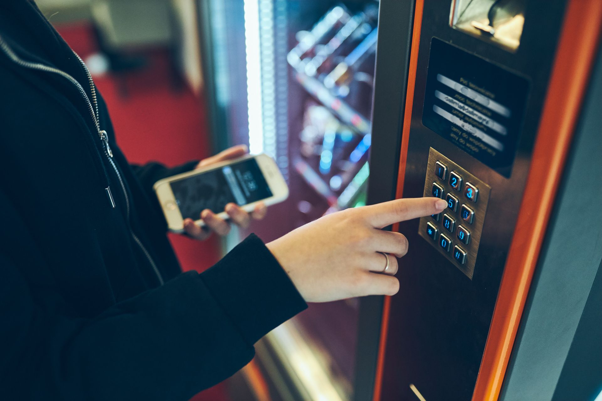 Person using a smartphone while pressing the keypad buttons on a vending machine
