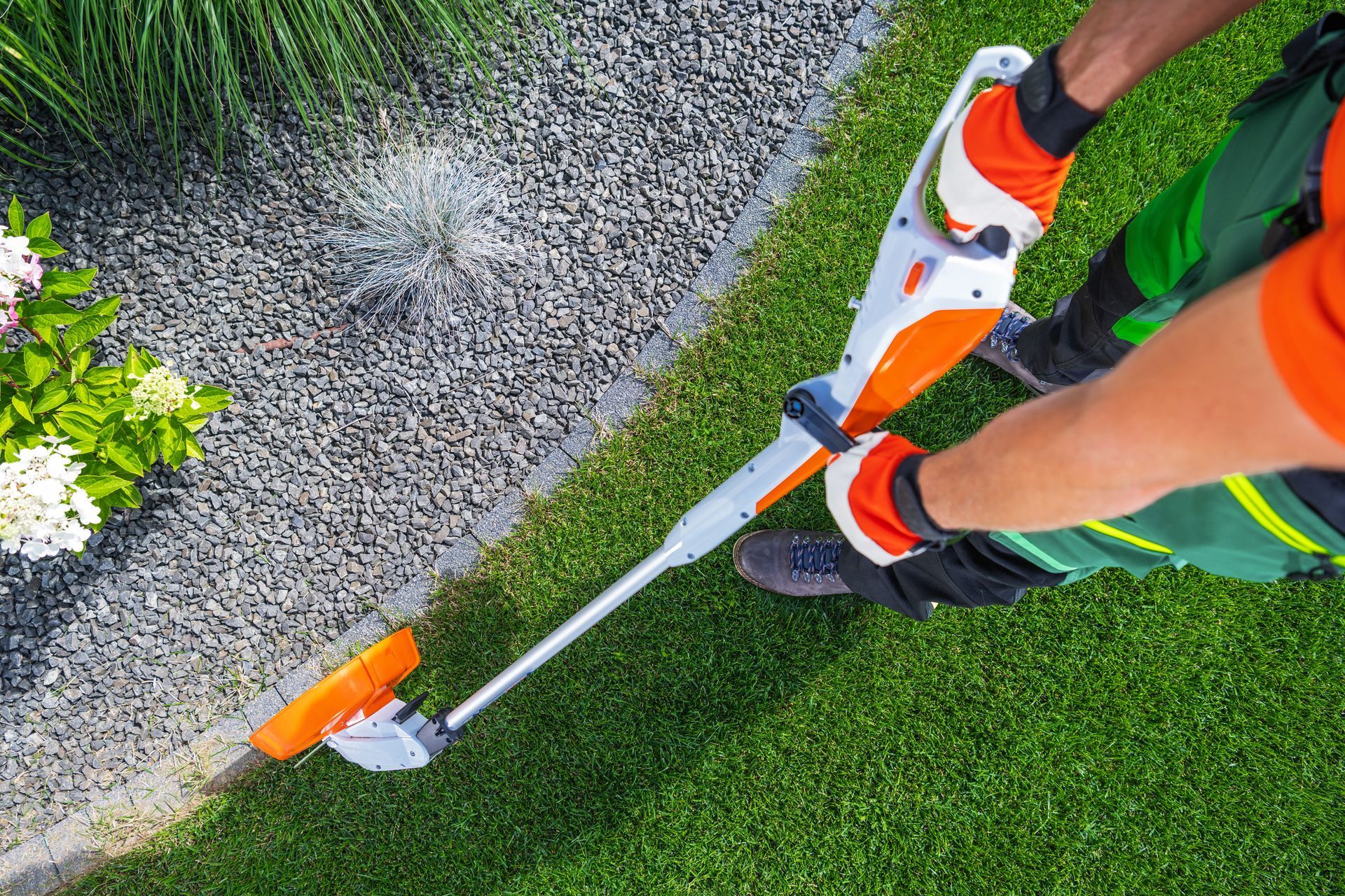 A person using a cordless orange and white string trimmer to edge a green lawn along a gravel path.