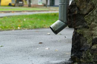 A drainpipe is attached to a stone wall on the side of the road.