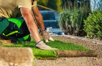 A man is kneeling down and laying a roll of grass in a garden.