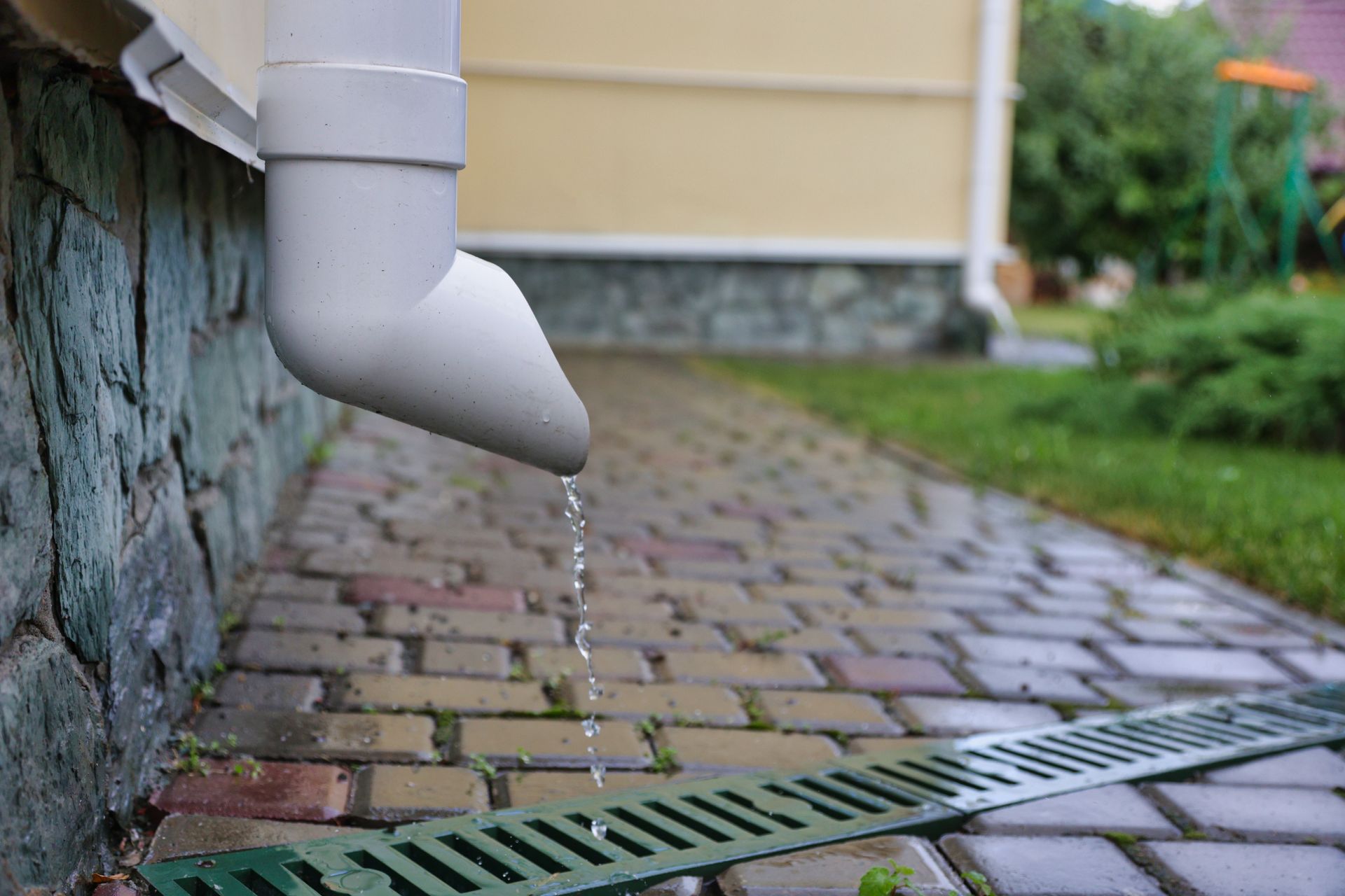 A drainpipe is leaking water on a sidewalk next to a house.
