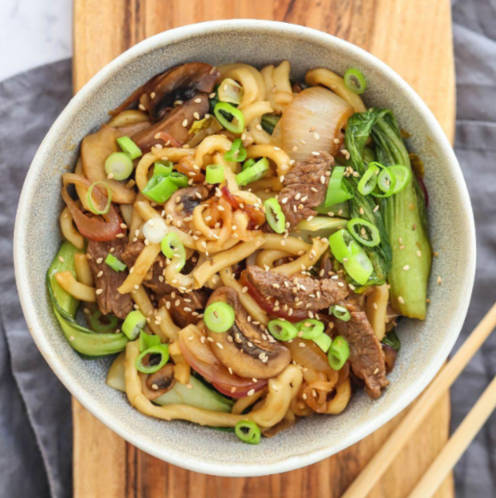 A bowl of noodles with meat and vegetables on a wooden cutting board with chopsticks.