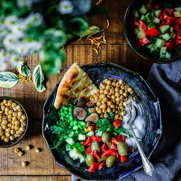 A bowl of food with chickpeas , vegetables and pita bread on a wooden table.