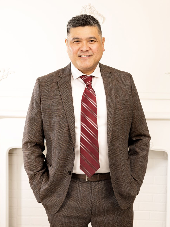 Man in brown suit, red striped tie, smiling, hands in pockets. Standing near a white fireplace.