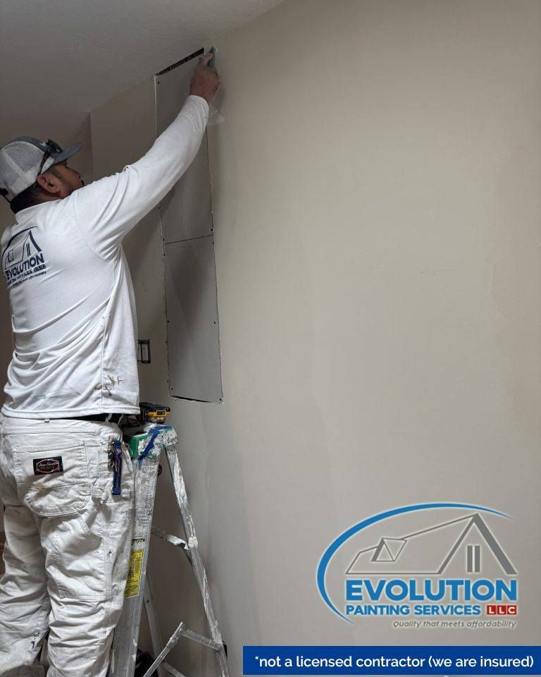 Painter on a ladder applying paint to a beige wall in an indoor room.