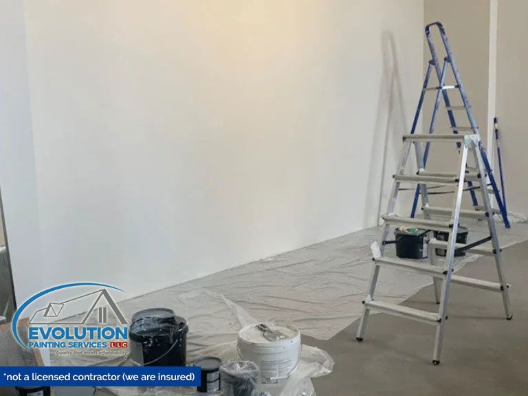 Painter’s room with white wall, covered floor, ladder, and paint buckets during renovation